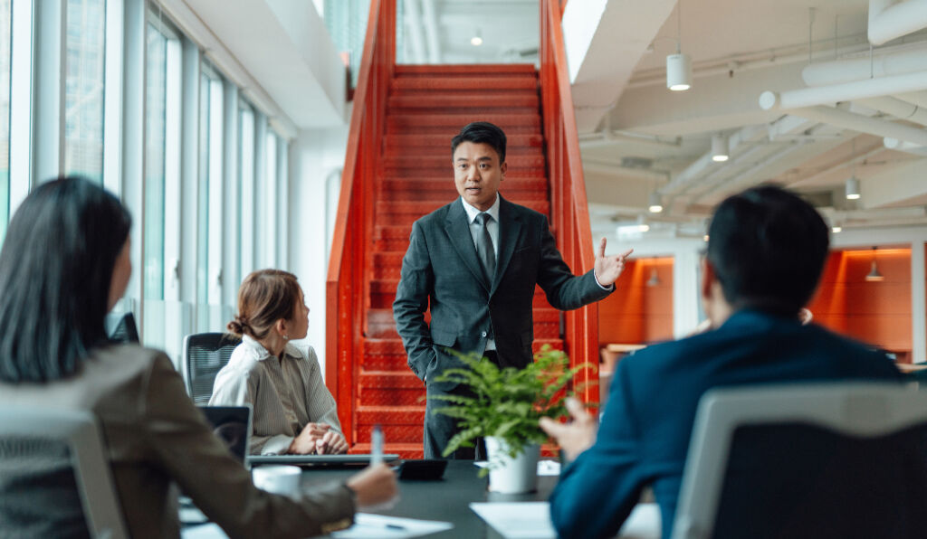 A man presenting in a modern meeting room, engaging with colleagues around a conference table.